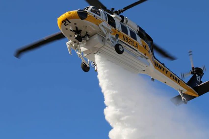 County fire Firehawk helicopter banking overhead mid-drop, water streaming from its belly tank against a clear sky